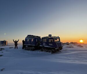 A tracked vehicle with two square cabins sits in the centre of the photo. It is parked on a snow covered ground, with the sun sitting just above the horizon to the right of frame. On the left of the photo is a man with his arms raised, and a small square hut. The sky is clear and being coloured by the setting sun.