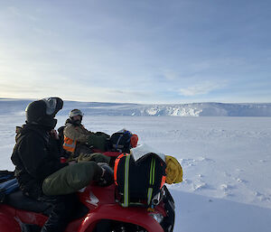 Two men on four wheel motorbikes are looking across a flat expanse of ice to the distance ice cliff that marks where the shoreline is.
