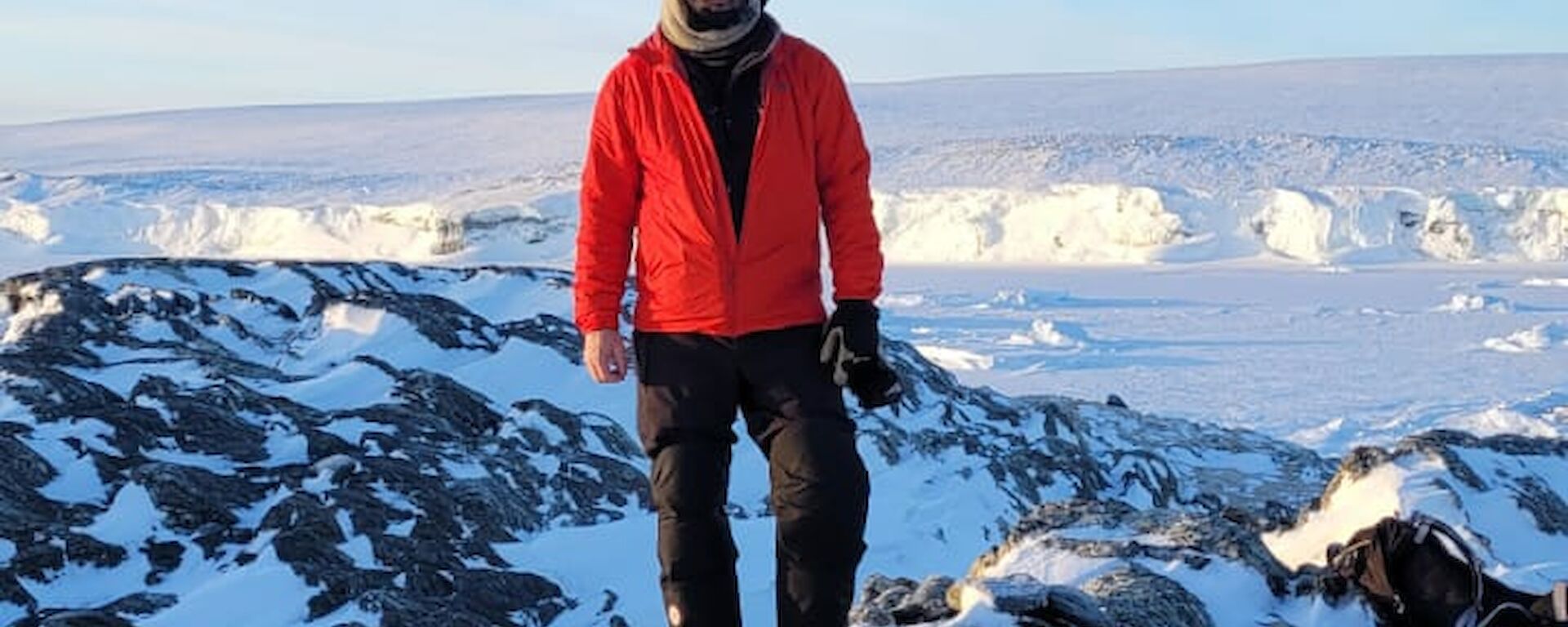 A man in a red jacket and beanie stands atop a snow covered rocky outcrop. In the distant background is the ice face of the shoreline. The sky is clear and blue.