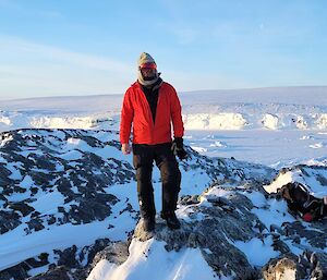 A man in a red jacket and beanie stands atop a snow covered rocky outcrop. In the distant background is the ice face of the shoreline. The sky is clear and blue.