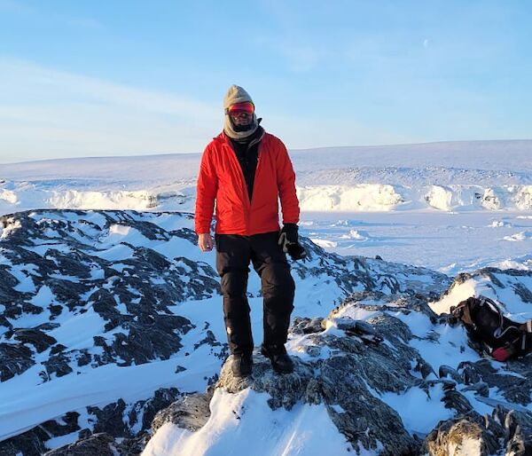 A man in a red jacket and beanie stands atop a snow covered rocky outcrop. In the distant background is the ice face of the shoreline. The sky is clear and blue.