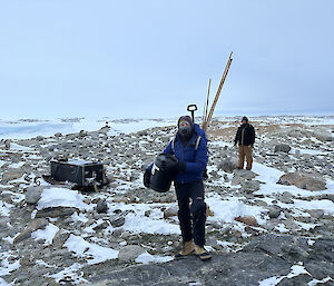 Two men are carrying supplies across a rocky hill top. The man in front has long bamboo canes, used to mark the location of survival stores.