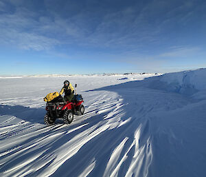 A man on a four wheel motorbike is stopped on a gently sloping snow covered stretch of ground, leading up off the sea ice behind him.