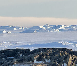 Icebergs are scattered in the distance, with a flat area of sea ice leading to a rocky hill in the foreground.