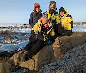 Four smiling people sit on a rocky outcrop with snow covered rocky hills visible in the background.