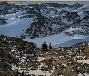 Two people walk down a rocky slope towards a snow-covered valley.