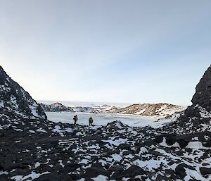 A rocky valley with two people visible in the distance near a frozen lake which is surrounded by snow covered hills.