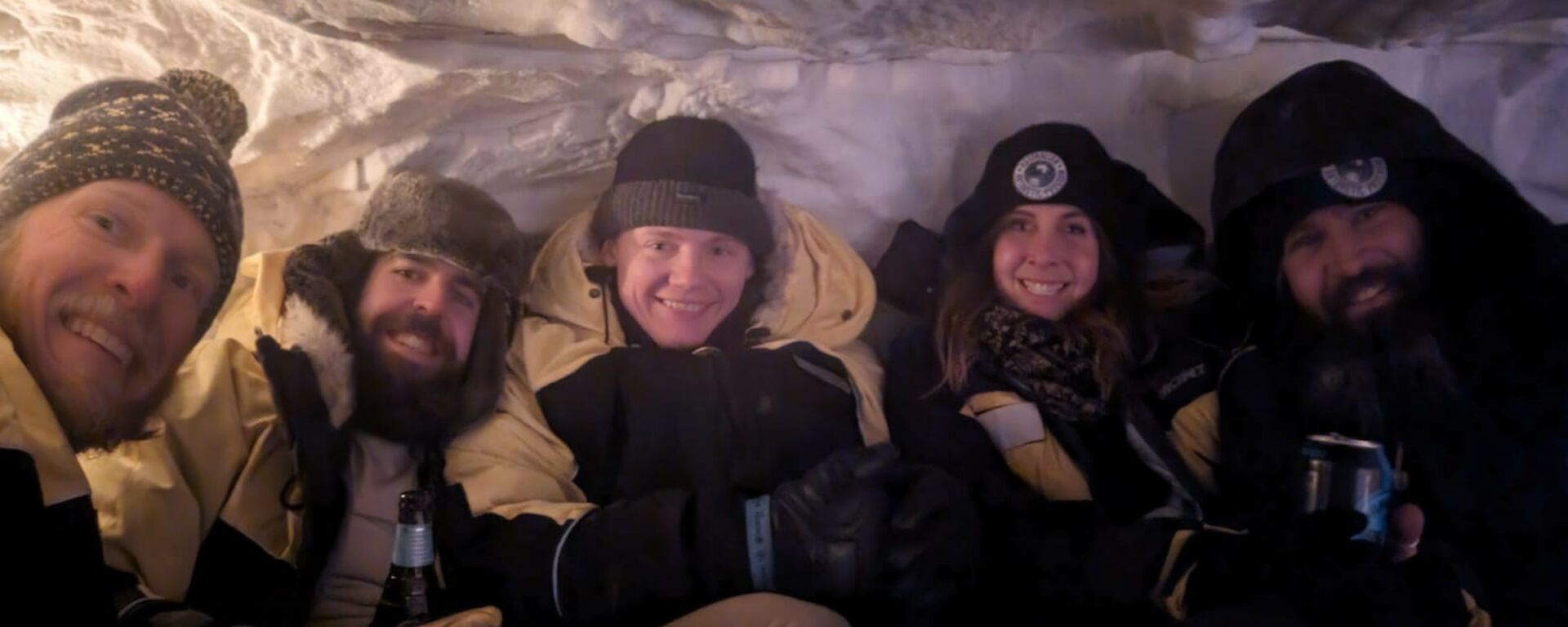 A group of five smiling people sitting inside a snow cave.