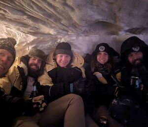 A group of five smiling people sitting inside a snow cave.