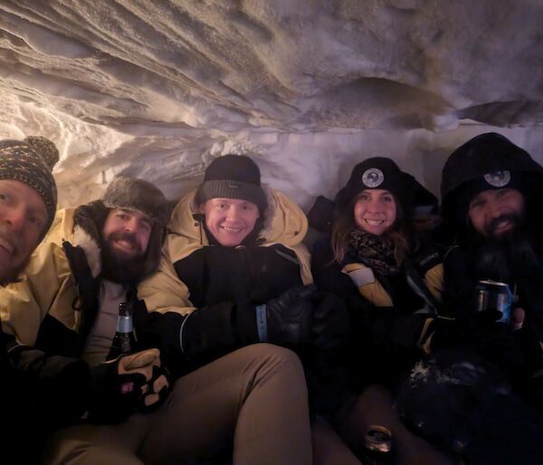 A group of five smiling people sitting inside a snow cave.