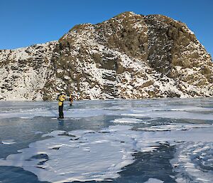Two men standing on a frozen lake partially covered in snow with a snow-covered rocky hill in the background.