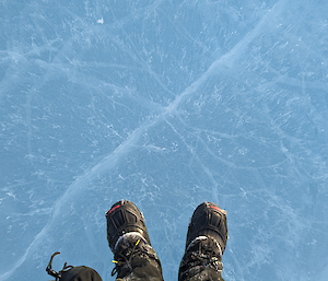 An overhead view of the frozen ice in clear-lake. The ice is a milky blue colour with cracks throughout. Two boots are visible at the bottomg of th ephotograph for scale.