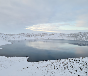 A large frozen lake partially covered by snow. The lake is surrounded by snow covered hills.