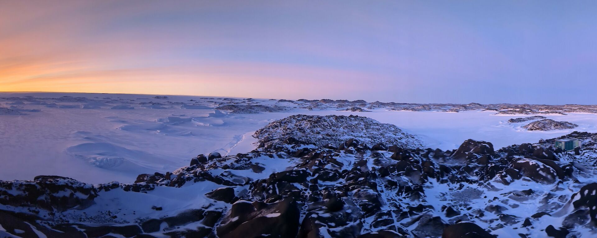 Panoramic photograph of the sun rising over the distant snow covered hills. Rocky outcrops are visible in the foreground.