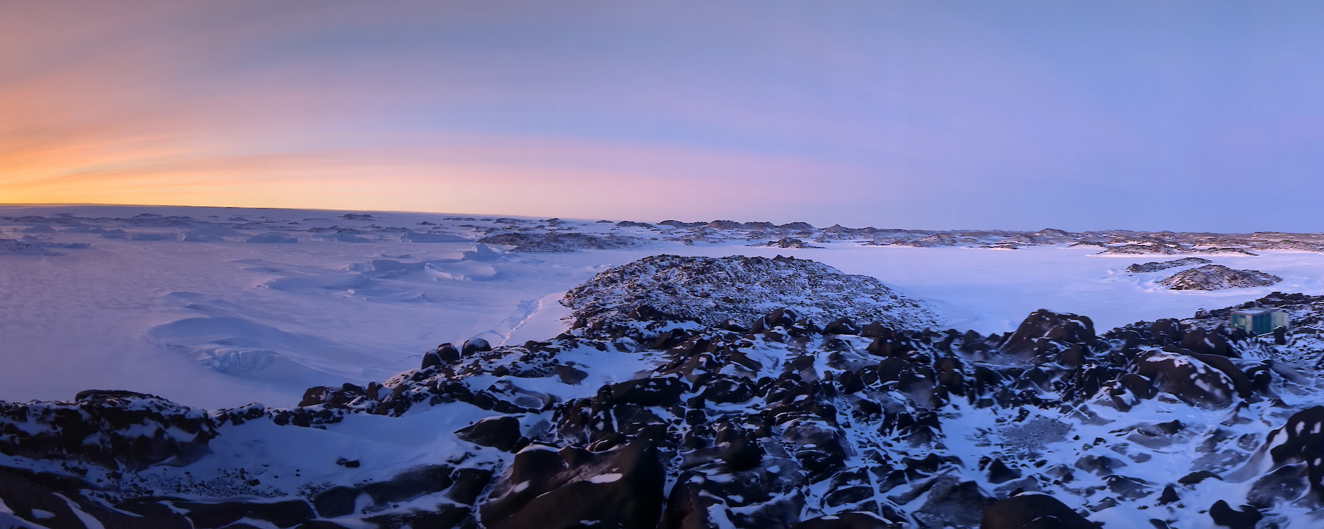 Panoramic photograph of the sun rising over the distant snow covered hills. Rocky outcorps are visible in the foreground.