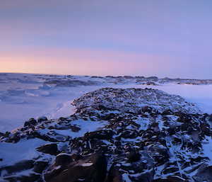 Panoramic photograph of the sun rising over the distant snow covered hills. Rocky outcorps are visible in the foreground.