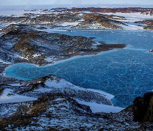 A frozen blue lake with numerous cracks throughout surrounded by snow-covered rocky hills.