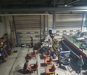 A large workshop is seen from above. There is a movie screen on the left side of the photo and a large tracked vehicle on the right with chairs setup on its rear deck. Theere are more chairs set up on the floor of the workshop.
