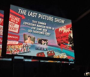 A introductory screen is visible on a movie screen in a large darkened room.