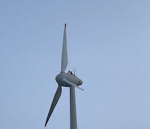 A man in a safety harness can be seen from the chest up at the top of a large wind turbine with three large blades.