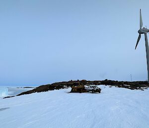 A large wind turbine stands on a rocky hill. There is a white utility vehicle and yellow dozer on the ice in front of it.