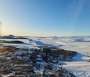 A number of brightly coloured buildings are visible on a rocky, snow and ice covered landscape. In the distance are rocky islands and two ice bergs frozen into sea ice