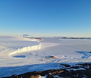 A photograph of an ice plateau on the left meeting a frozen sea on the right. In the foreground is a rocky hillside.