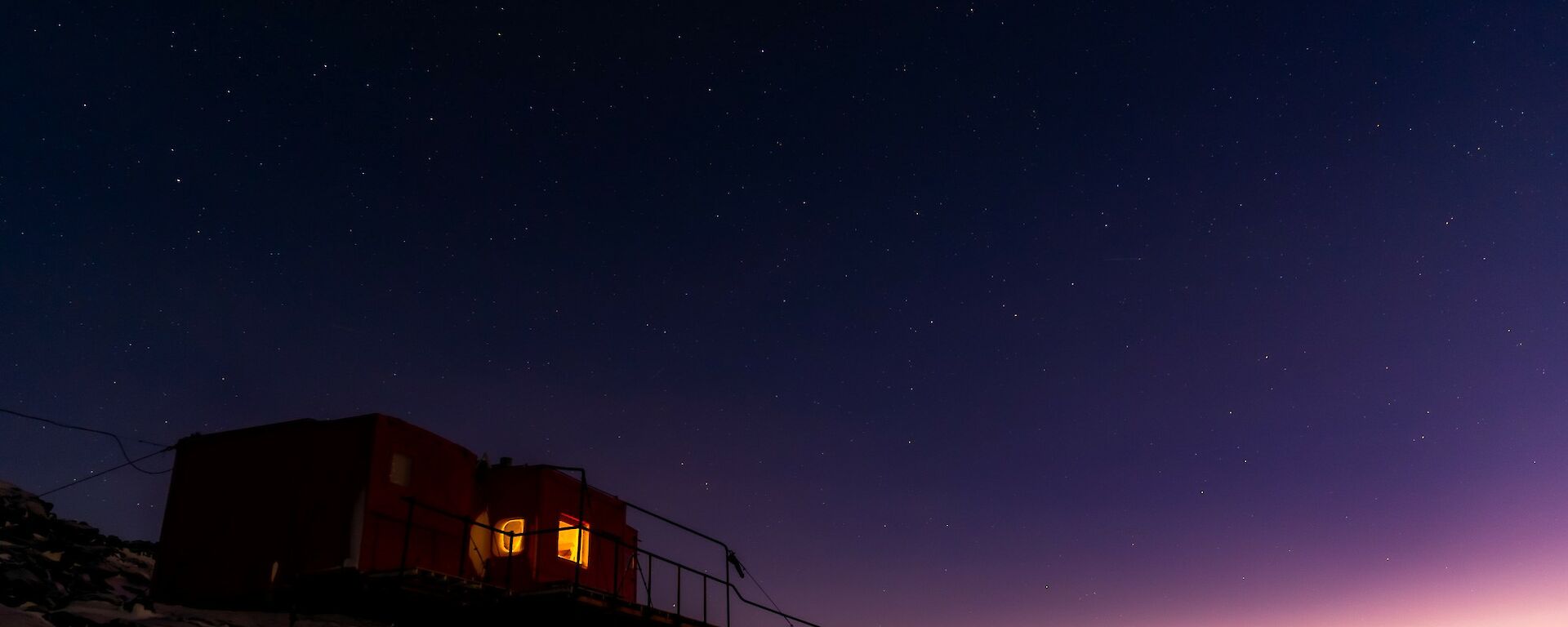 A red field hut sits on a snowy rocky outcrop with the stars and night sky visible above it.