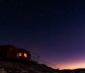 A red field hut sits on a snowy rocky outcrop with the stars and night sky visible above it.