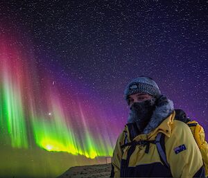 A man stands outside a building under a night sky with a brilliant curtain of an aurora in vivid yellow, green and pink.