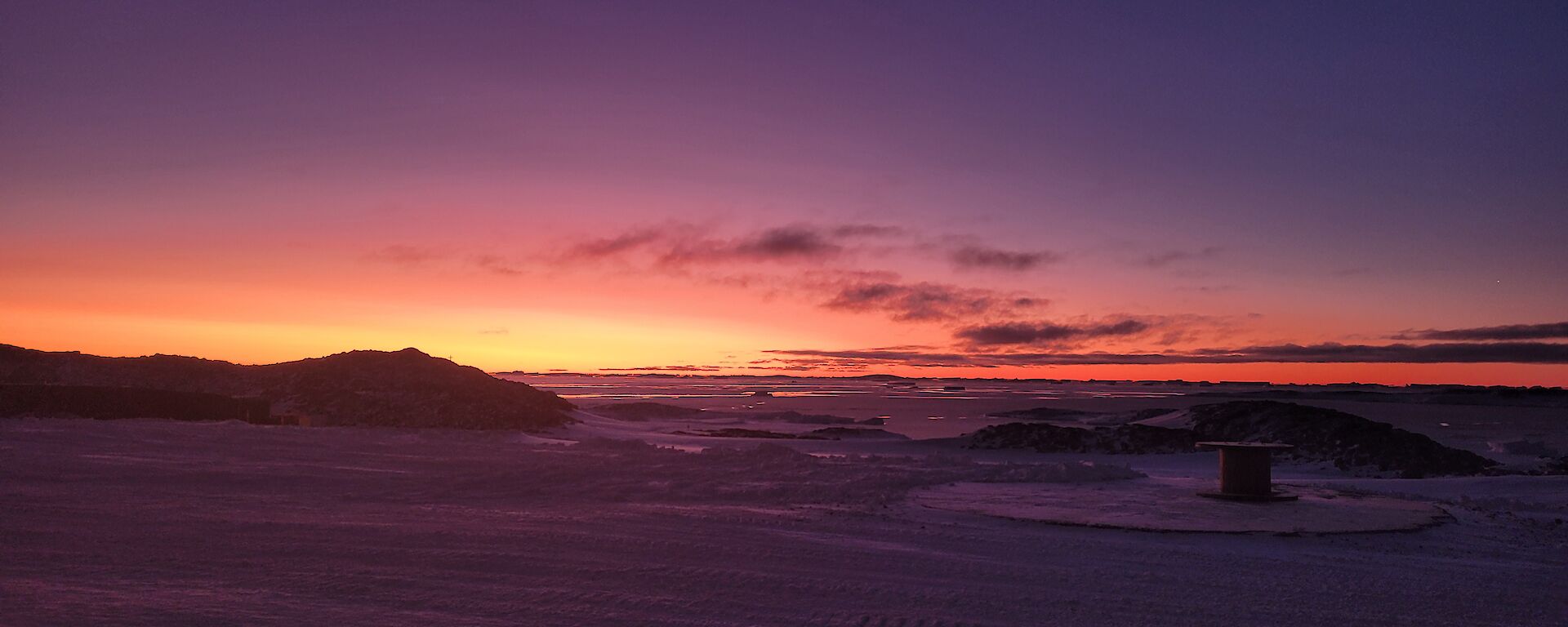 This view is looking across the snow covered ground from station. There is low rocky hills in the middle of the photo, with the sky lit up with colours from the sun which has set below the horizon.