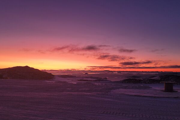 This view is looking across the snow covered ground from station. There is low rocky hills in the middle of the photo, with the sky lit up with colours from the sun which has set below the horizon.