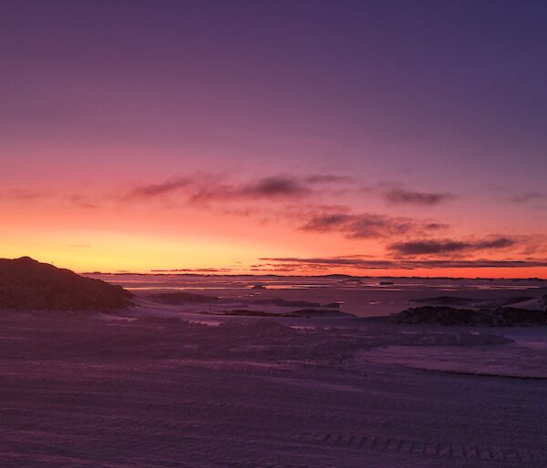 This view is looking across the snow covered ground from station. There is low rocky hills in the middle of the photo, with the sky lit up with colours from the sun which has set below the horizon.