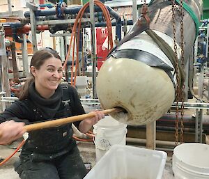 A woman is kneeling in front of a long white metal cylinder that is supported in the air by a sling. She is digging material out of a hole in the end of the cylinder with a small shovel. There is a maze of pipework in the background.