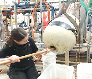 A woman is kneeling in front of a long white metal cylinder that is supported in the air by a sling. She is digging material out of a hole in the end of the cylinder with a small shovel. There is a maze of pipework in the background.