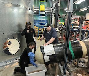 A woman is kneeling in front of a long metal cylinder that is supported in the air by a sling. She is digging material out of a hole in the end of the cylinder with a small shovel. There is a maze of pipework in the background, and a large metal water tank to the left.