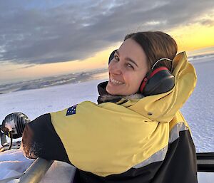 A woman in a big yellow cold weather jacket is standing in the door arch of a Hagglund vehicle. She is looking back at the camera and smiling. The view in front is of an expanse of snow, continuing down to the ice covered water in front of Casey Station. The sky is coloured by the sunset.