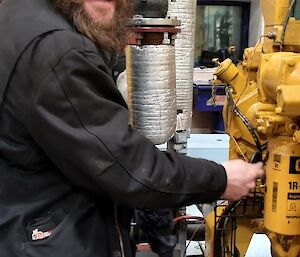 A man wearing hearing protection is working on a large yellow engine. He is looking at the camera and smiling.