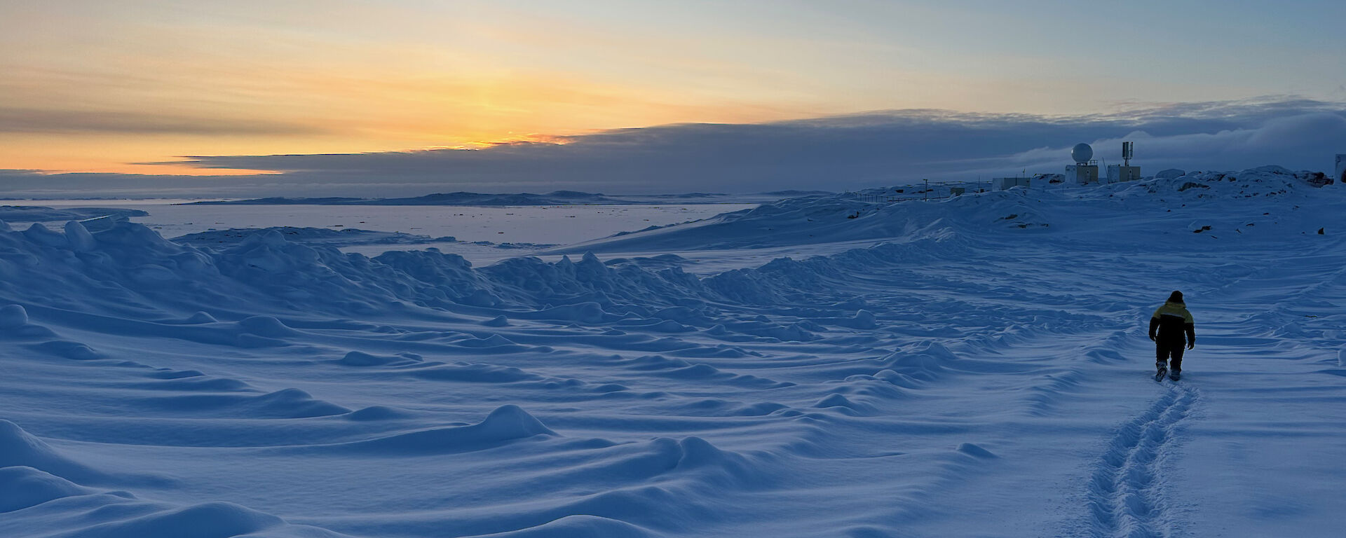 A person walks aways from camera leaving fresh footprints in the snow under a moody sky