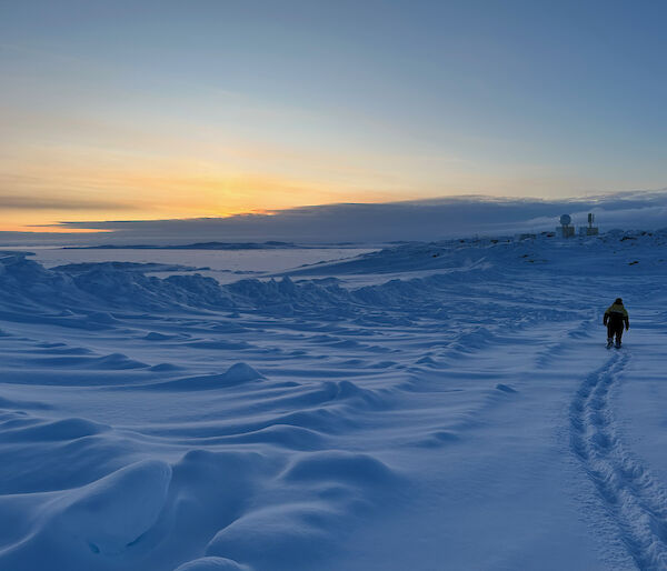 A person walks aways from camera leaving fresh footprints in the snow under a moody sky