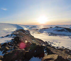 The sun is setting in the distance with a rocky ridgeline leading towards it. On the left is a large glacier and in the valley to the right and towards the horizon are a large number of penguins.