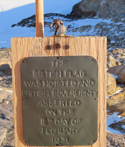 A small figurine is on top of a wooden board with a plaque proclaiming sovereignty. In the background is a rocky, snow and ice covereed landscape.