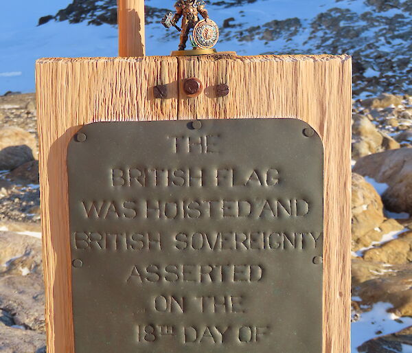 A small figurine is on top of a wooden board with a plaque proclaiming sovereignty. In the background is a rocky, snow and ice covereed landscape.