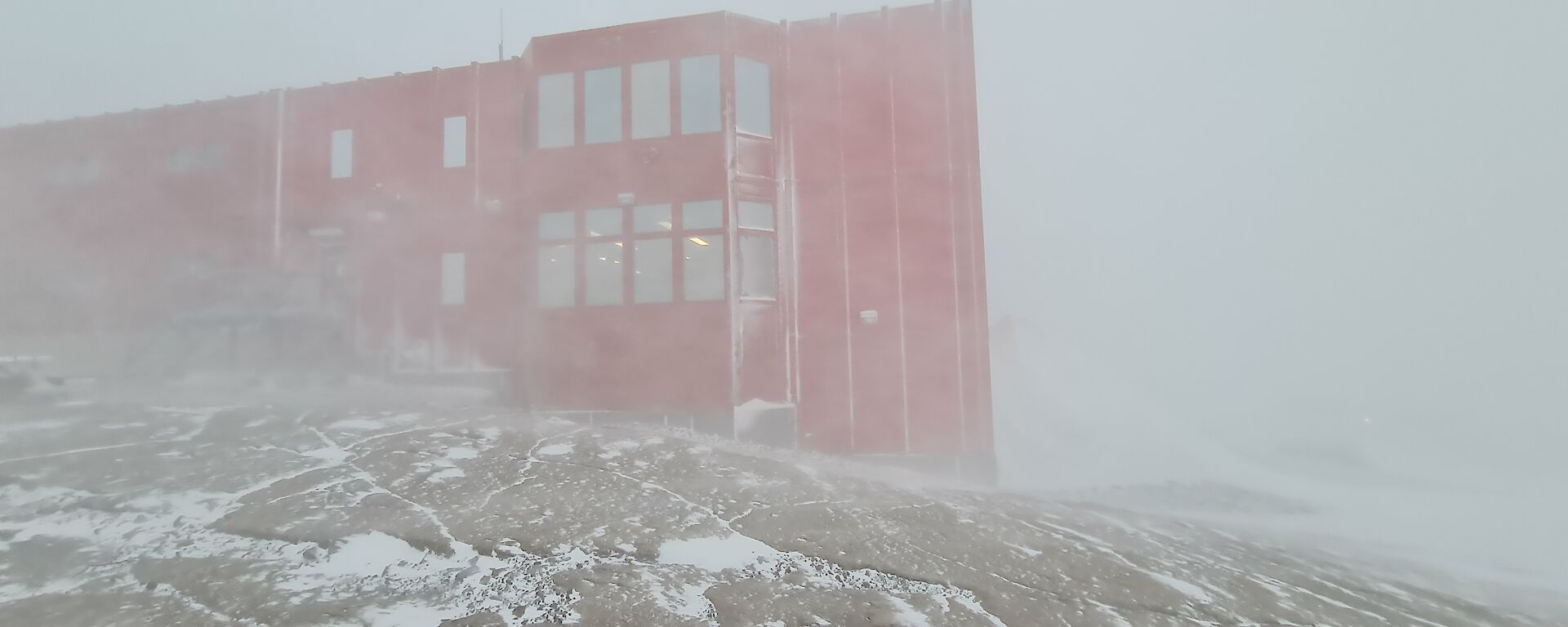 A large red building on a rocky, snow covered slope is being obscured by the beginning of a snow blizzard.