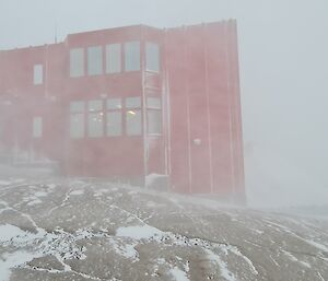 A large red building on a rocky, snow covered slope is being obscured by the beginning of a snow blizzard.