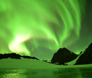 A very bright green aurora is in the night sky above a rocky, ice covered landscape. There is an ice lake in the foreground that is reflecting the aurora.