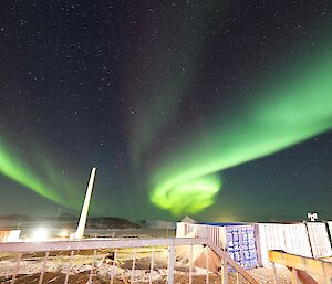 A bright green and purple aurora is visible above a rocky landscape. There are shipping containers to the bottom right of frame and a tall tower to the left of frame.