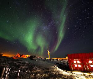 A green and purple aurora is visible in the night sky around a wind turbine. There is a red building with lit windows in the bottom right of fram