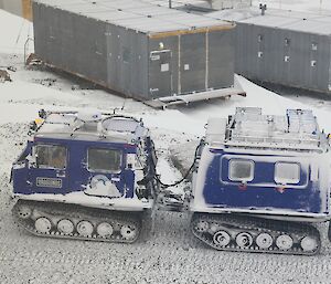 A blue Hägglunds vehicle is driving on a snow covered track. There are old grey buildings in the background.