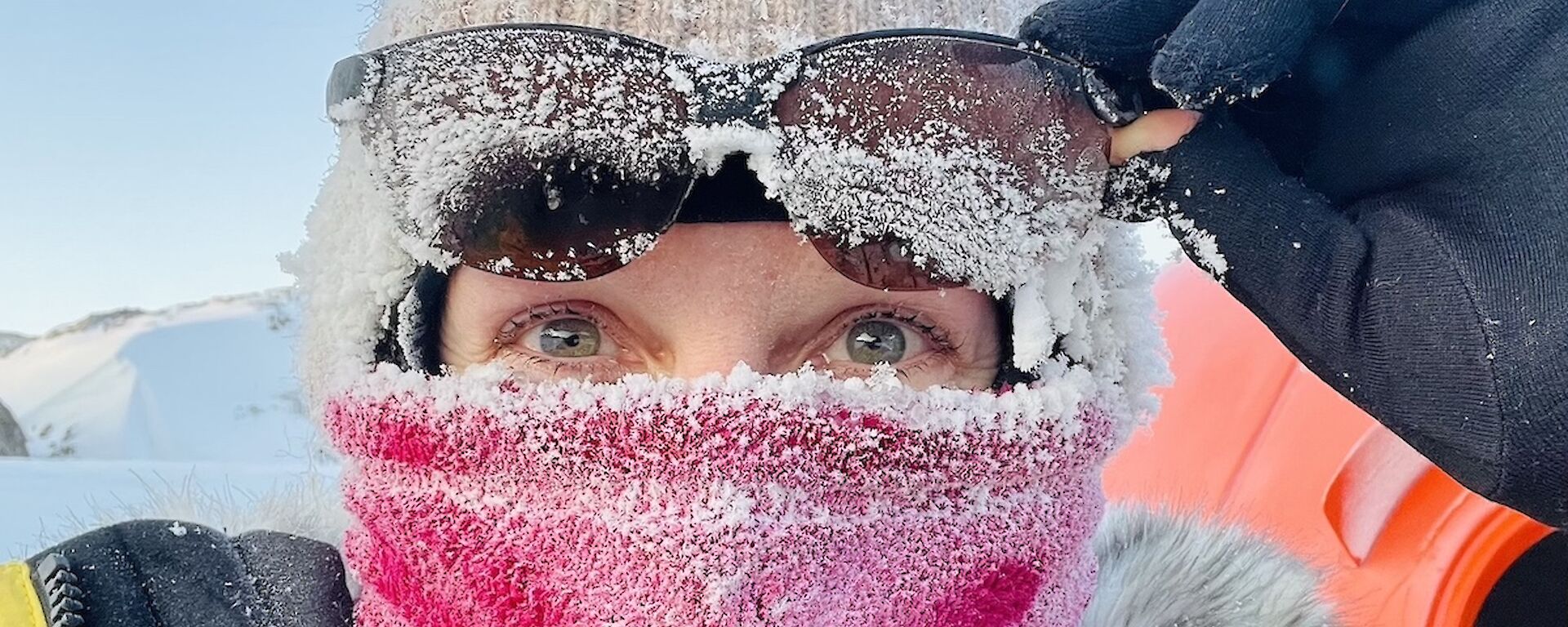 A lady wears a pink face buff, beanie and sunglasses perched on her head, all covered in a thick layer of ice crystals.
